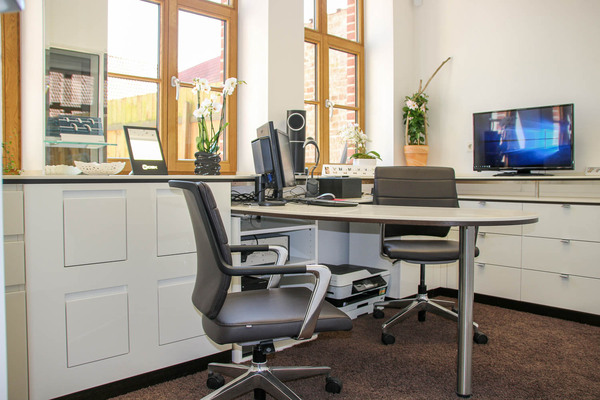 Modernes Büro mit Schreibtisch, Computer, Drehstühlen und großen Fenstern im Tageslicht.